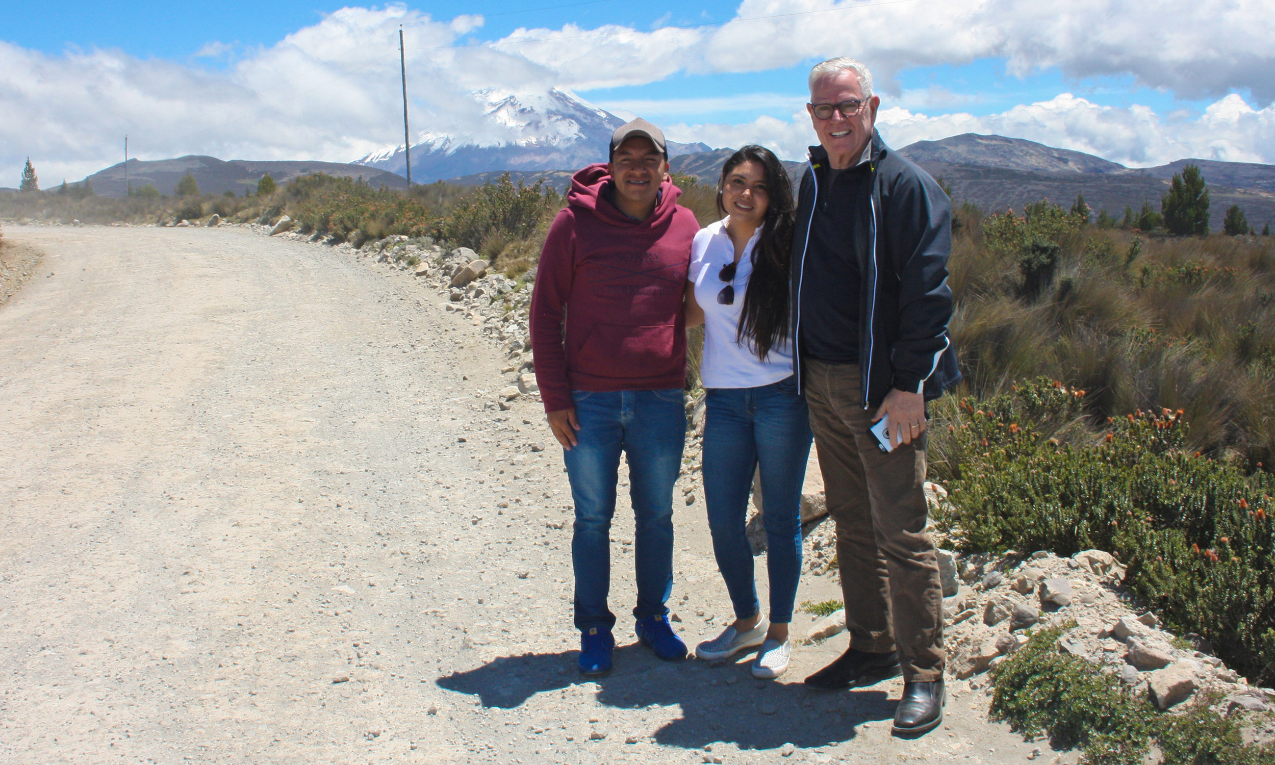 Three people stand to the side of a dirt road with blue skies