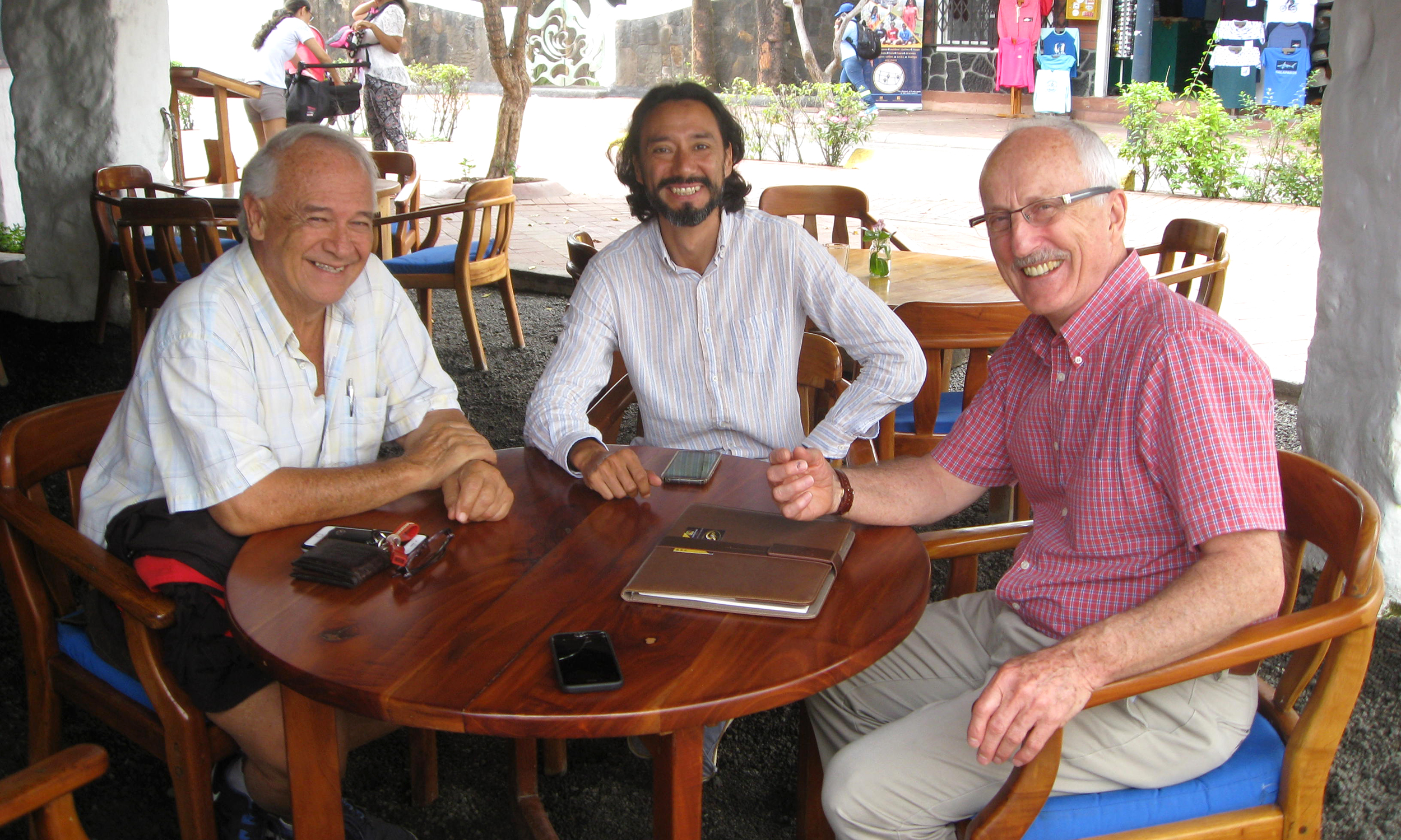 Three men in Ecuador sit at a wooden table at wooden chairs and smile at camera